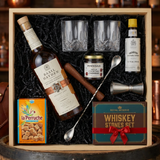 Basil Hayden's Old-Fashioned cocktail gift crate with Basil Hayden's Kentucky Straight Bourbon Whiskey, crystal glasses, bitters, muddler, bar spoon, jigger, cocktail cherries, sugar cubes, and whiskey stones nestled in black zigzag crinkle paper inside wooden crate on rustic aged oak surface with warm amber bourbon bar lighting