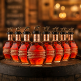 Blanton's Gold Edition Single Barrel Bourbon complete 8-bottle collector's set with B-L-A-N-T-O-N-S gold horse and jockey stoppers, 750ml bottles arranged in row on rustic aged oak barrel with warm amber bourbon bar lighting and moody bokeh