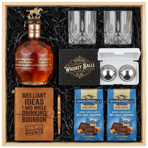 Blanton's Gold Edition Single Barrel Bourbon gift crate with gold horse stopper, crystal-cut old fashioned glasses, faux-leather bourbon journal and pen set, premium XXL stainless steel whiskey balls with freezer tray, and two Ghirardelli Dark Chocolate Sea Salt Caramel bags in black crinkle paper on white background
