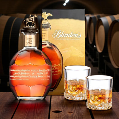 Blanton’s Gold Edition Single Barrel Bourbon Gift Set featuring 750ml bottle, gold box, and two glasses – displayed in a wooden barrel room scene – exclusive collector’s bourbon from Rare Bourbon Shop