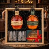 Blanton's Original Single Barrel Bourbon 750ml and Blanton's Special Reserve Green Label 700ml dual-bottle gift crate with bronze horse stoppers, crystal-cut old fashioned glasses, and Royal Reserve whiskey stones gift box with red ribbon nestled in black crinkle paper inside wooden crate on rustic aged oak surface with warm amber bourbon bar lighting and moody bokeh