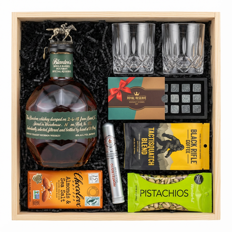 Blanton's Special Reserve Green Label Single Barrel Bourbon 700ml gift crate with bronze horse stopper and green label with white text, crystal-cut old fashioned glasses, Royal Reserve whiskey stones set with tray, Montecristo Platinum cigar tube, Black Rifle Coffee Tactisquatch blend, Chocolove Almonds & Sea Salt dark chocolate bar, and Wonderful Pistachios in black crinkle paper on white background