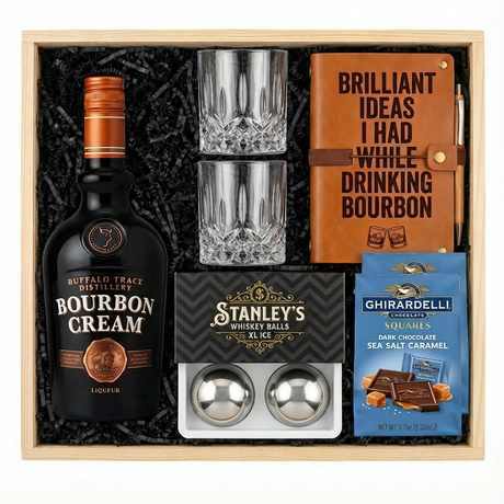 Buffalo Trace Bourbon Cream Liqueur gift crate with black bottle and copper cap, crystal-cut old fashioned glasses, tan leather bourbon journal and pen set, Stanley's XXL stainless steel whiskey balls with rectangular freezer tray, and two Ghirardelli Dark Chocolate Sea Salt Caramel gable-top bags layered to show both all nestled in black zigzag crinkle paper inside wooden crate on white background