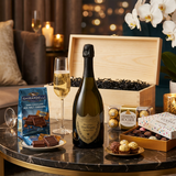 Dom Pérignon Vintage 2015 Champagne gift crate lifestyle scene with bottle, two champagne flutes with golden bubbly pour, Ghirardelli Dark Chocolate Sea Salt Caramel squares, Ferrero Rocher chocolates, Godiva Goldmark Let's Celebrate assorted chocolates, and open wooden crate on elegant marble surface in luxurious celebration setting with warm ambient lighting.