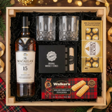 Bird's eye view of The Macallan 15 Year Old Double Cask Single Malt Scotch gift crate with crystal-cut old fashioned glasses, premium whiskey stones gift set, Ferrero Rocher chocolates, and Walker's shortbread fingers on elegant dark wood surface with warm golden bokeh lights and holiday decor