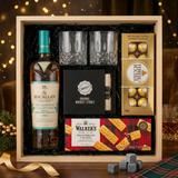 Bird's eye view of The Macallan Harmony Collection Phoenix Honey Orchid Tea Jing gift crate with crystal-cut old fashioned glasses, premium whiskey stones gift set, Ferrero Rocher chocolates, and Walker's shortbread fingers on elegant dark wood surface with warm golden bokeh lights and holiday decor
