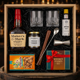 Maker's Mark Old-Fashioned cocktail gift crate with Maker's Mark bourbon with red wax seal, crystal glasses, bitters, muddler, bar spoon, jigger, cocktail cherries, sugar cubes, and whiskey stones nestled in black zigzag crinkle paper inside wooden crate on rustic aged oak surface with warm amber bourbon bar lighting
