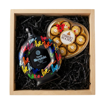 Mándala Love Añejo Tequila gift crate with black ceramic decanter featuring colorful love script and heart-shaped Ferrero Rocher hazelnut chocolate box in natural wood crate with black crinkle fill, romantic gift