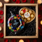 Bird's eye view of gift crate with Mándala Love Añejo Tequila bottle featuring colorful love graffiti and heart-shaped Ferrero Rocher chocolate box nestled in black crinkle paper with red rose petals