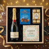Moët & Chandon Impérial Brut Champagne gift crate with gold foil neck and red wax seal, Ghirardelli Dark Chocolate Sea Salt Caramel squares, Godiva Let's Celebrate chocolate box, and Ferrero Rocher chocolates on elegant dark wood surface with warm golden bokeh lights and celebratory decor