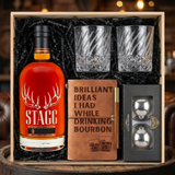Stagg Kentucky Straight Bourbon Whiskey barrel proof gift crate with antler logo bottle, twisted crystal-cut old fashioned glasses, tan leather bourbon journal and pen set, and Stanley's XXL stainless steel whiskey balls realistically nestled in black zigzag crinkle paper inside wooden crate on rustic aged oak surface with warm amber bourbon bar lighting and moody bokeh