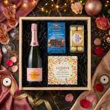 Veuve Clicquot Rosé Brut Champagne gift crate with Ghirardelli Dark Chocolate Sea Salt Caramel squares, Godiva Let's Celebrate chocolate box, and Ferrero Rocher chocolates on elegant surface with warm golden bokeh lights and rose gold celebratory decor